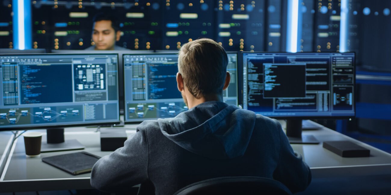 IT Specialist Works on Personal Computer with Screens Showing Software Program with Coding Language Interface. In the Background Technical Room of Data Center with Professional Working