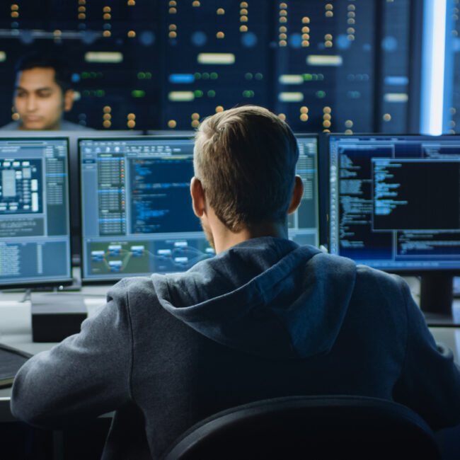 IT Specialist Works on Personal Computer with Screens Showing Software Program with Coding Language Interface. In the Background Technical Room of Data Center with Professional Working